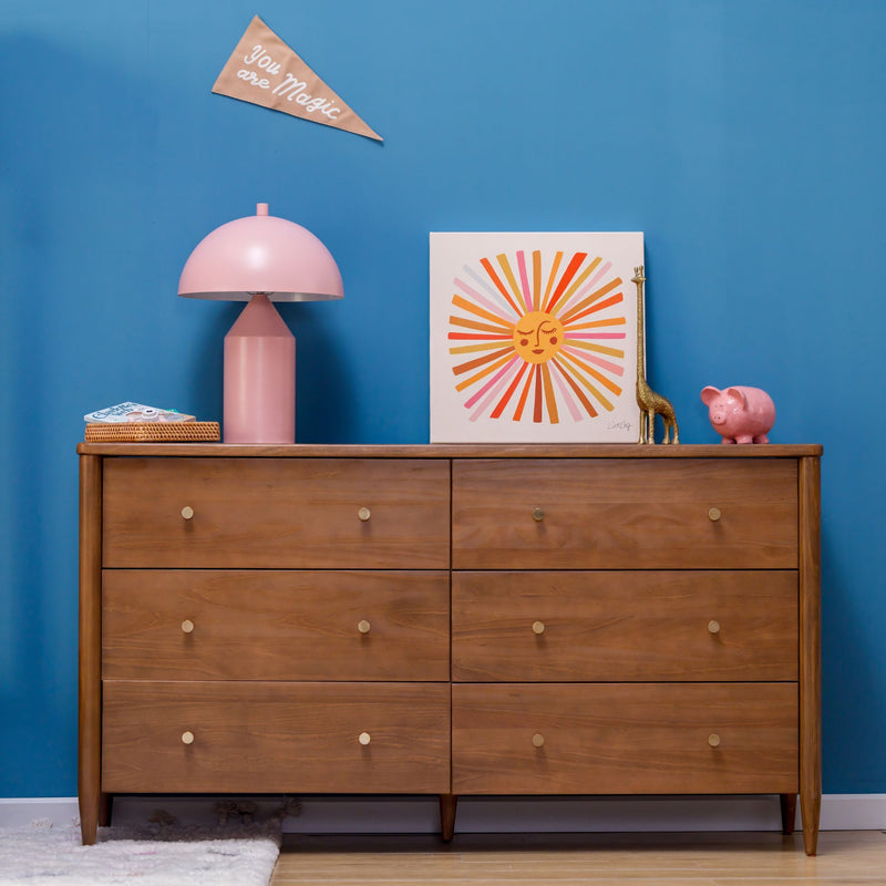 Six-drawer mid-century modern wooden dresser against a blue accent wall with a pink mushroom lamp, sunburst art, gold giraffe figurine and pink piggy bank, nursery decor