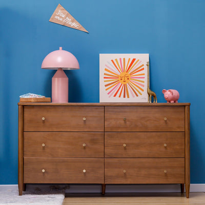 Six-drawer mid-century modern wooden dresser against a blue accent wall with a pink mushroom lamp, sunburst art, gold giraffe figurine and pink piggy bank, nursery decor