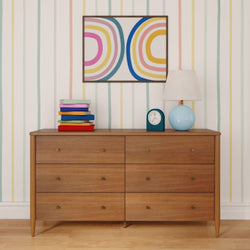 Mid-century modern wooden six-drawer dresser with colorful stacked books, blue table lamp, teal alarm clock and abstract rainbow wall art on pastel striped wallpaper