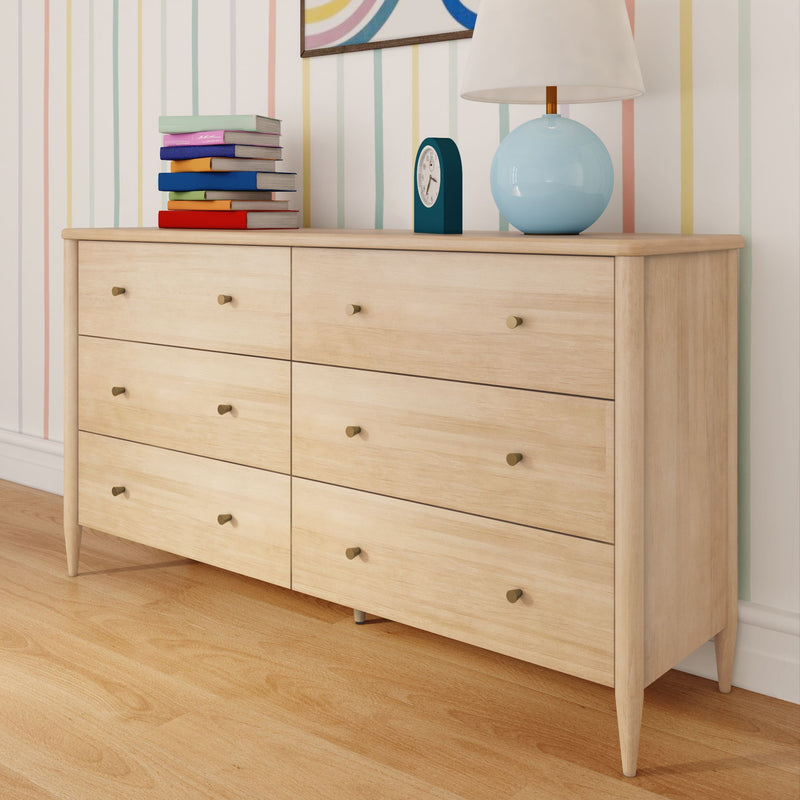 Light wood six-drawer dresser on hardwood floor, modern mid-century storage cabinet topped with a stack of colorful children's books, teal alarm clock and blue ceramic lamp against a pastel-striped wall