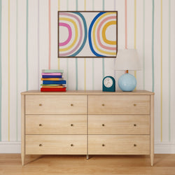Light wood six-drawer dresser topped with a blue globe table lamp, teal alarm clock, stack of colorful books, and framed retro rainbow art against pastel striped wallpaper