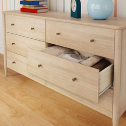 Scandinavian light wood six-drawer bedroom dresser with an open drawer revealing folded linens, brass knobs, books, clock and lamp on top