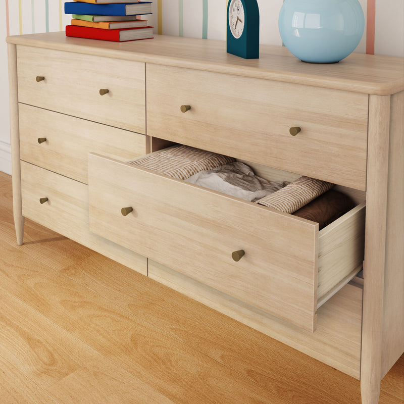 Scandinavian light wood six-drawer bedroom dresser with an open drawer revealing folded linens, brass knobs, books, clock and lamp on top
