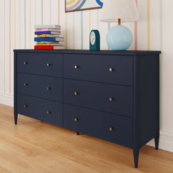 Mid-century modern navy blue six-drawer dresser with brass knobs on hardwood floor in a pastel-striped nursery, topped with a stack of colorful children’s books, teal alarm clock and light blue table lamp