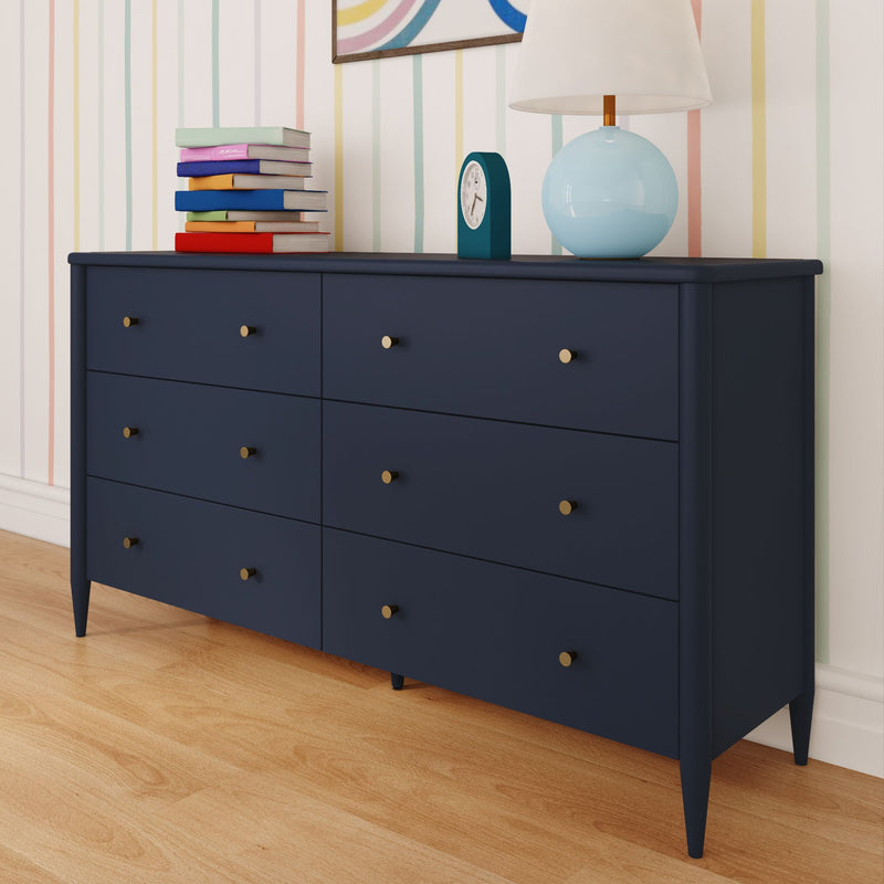 Mid-century modern navy blue six-drawer dresser with brass knobs on hardwood floor in a pastel-striped nursery, topped with a stack of colorful children’s books, teal alarm clock and light blue table lamp