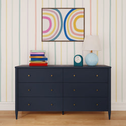Navy blue six-drawer dresser in a kids' room with colorful striped wallpaper, stack of children's books, blue table lamp, retro alarm clock and abstract rainbow wall art