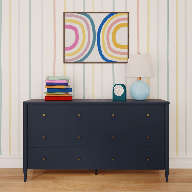 Navy blue six-drawer dresser in a kids' room with colorful striped wallpaper, stack of children's books, blue table lamp, retro alarm clock and abstract rainbow wall art