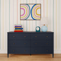 Navy blue six-drawer dresser against pastel striped wallpaper with abstract rainbow wall art, blue table lamp, teal clock and stack of colorful books on wooden floor