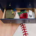 Open navy dresser drawer filled with neatly folded children's shirts and pajamas beside a baseball-stitched round rug on light hardwood floor