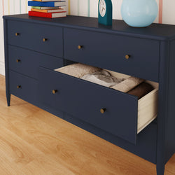 Modern navy blue six-drawer dresser with brass knobs and a partially open drawer showing folded blankets and linens, styled on hardwood floor with stacked books, teal clock, and blue table lamp on top