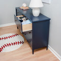 Navy blue dresser with open drawer full of children’s clothes beside a white lamp and baseball rug on hardwood floor