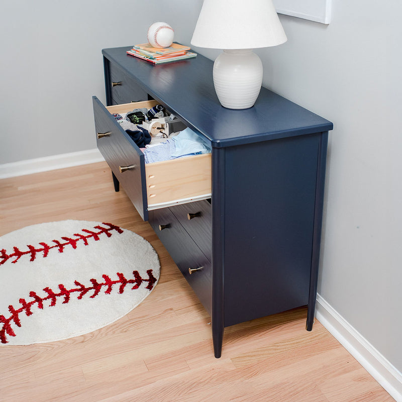 Navy blue dresser with open drawer full of children’s clothes beside a white lamp and baseball rug on hardwood floor