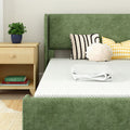 Modern green upholstered bed with white mattress and folded towel, wooden nightstand with potted plant and books, decorative striped and gingham pillows