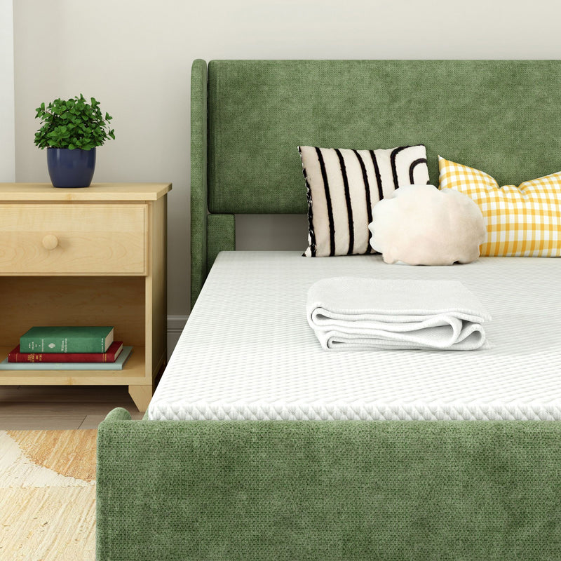 Modern green upholstered bed with white mattress and folded towel, wooden nightstand with potted plant and books, decorative striped and gingham pillows