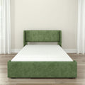 Green upholstered platform bed with padded headboard and white mattress in a modern minimalist bedroom with hardwood floors, neutral walls, and sheer curtains