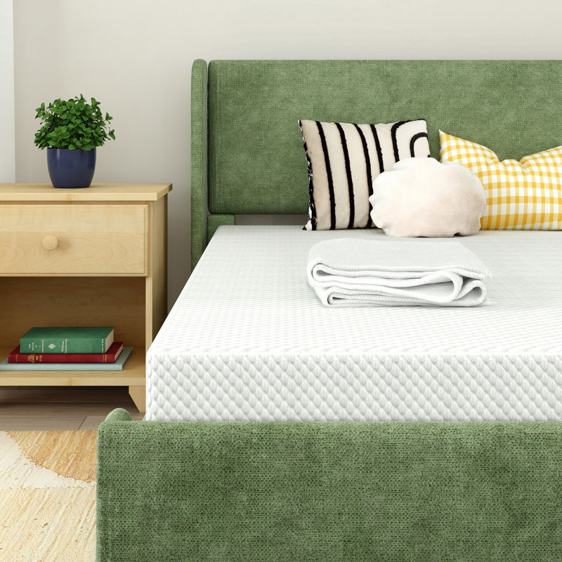 Modern bedroom with green upholstered bed and headboard, white quilted mattress and folded blanket, striped and yellow gingham decorative pillows, wooden nightstand with potted plant and books.