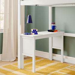 White kids’ study desk tucked under a loft bed with a blue desk lamp, stacked puzzle cube, mug of colorful markers and books against a green wall and yellow striped rug
