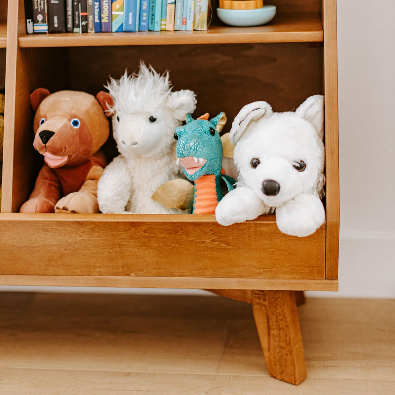 Children's stuffed animals on a mid-century wooden shelf — brown bear, white sheep, teal dragon and white husky plush beneath children's books in a bright nursery playroom