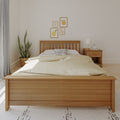 Minimalist neutral bedroom with wooden slat bed frame, beige linen bedding and spotted accent pillow, wooden nightstand with lamp and alarm clock, framed fruit prints and potted snake plant, modern Scandinavian decor