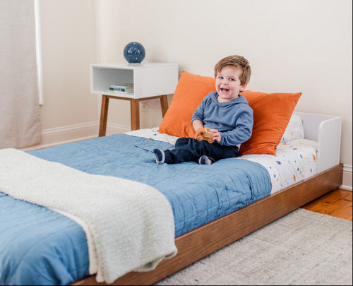 Child sitting on a bed with blue bedding and orange pillows in a room.