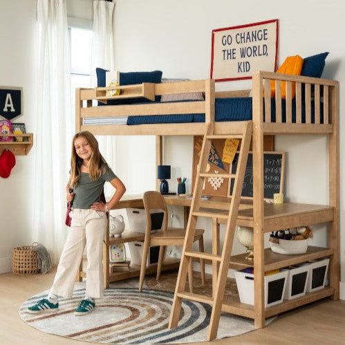 Natural wood kids loft bed with built-in desk, ladder and storage bins in a modern children's bedroom, round rug, wall art reading "Go change the world, kid" and a smiling girl.