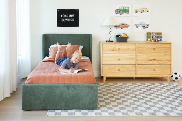 Young boy reading on a green upholstered bed with orange quilt in a modern kids bedroom featuring wooden dresser, toy cars, checkered rug and "Long Live Boyhood" wall sign