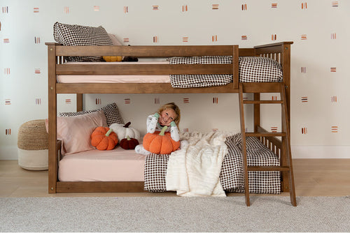 A wooden bunk bed with a child sitting underneath on a light gray carpeted floor against a patterned wallpaper background. The bed is styled with pink bedding, black and white gingham pillows, a cream knit blanket, and orange pumpkin plush toys.