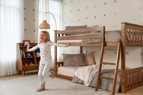 A child playing in a bright room with a wooden low loft bed with checkered print bedding and a toy shelf in the background.