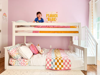 Kids loft bunk bed in pastel pink bedroom with smiling child, white bunk bed, colorful checkered quilt, heart-pattern sheets, unicorn plush and "Flower Babe" wall art